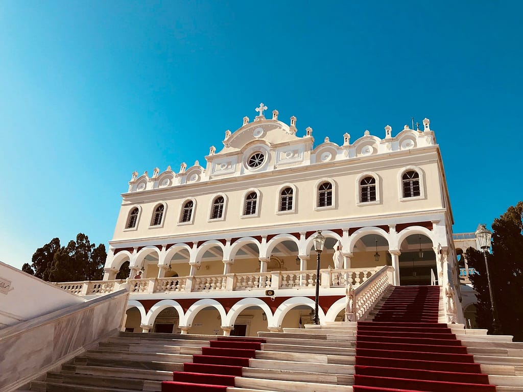 a large white building with red stairs with Tinos in the background
